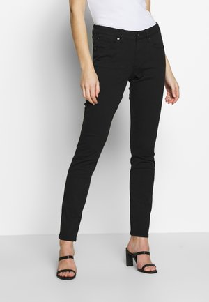 Femme portant un pantalon noir slim, un haut blanc et des sandales noires à talons hauts, debout devant un fond clair.