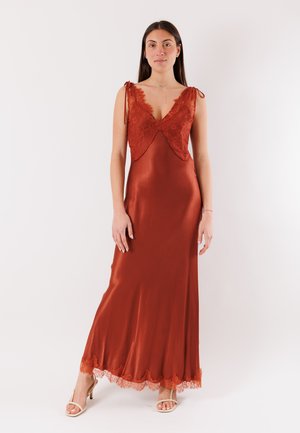 Donna in piedi che indossa un abito senza maniche color ruggine in satin con dettagli in pizzo sulla parte superiore e sull'orlo, abbinato a tacchi beige aperti sulla punta.