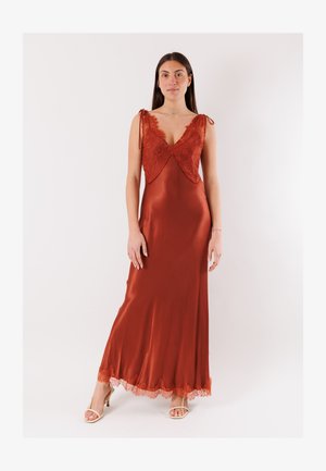 Donna in piedi che indossa un abito senza maniche color ruggine in satin con dettagli in pizzo sulla parte superiore e sull'orlo, abbinato a tacchi beige aperti sulla punta.