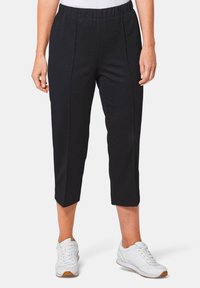 Zwarte cropped broek met een elastische tailleband, voorzien van verticale naden. Gedragen met witte sportschoenen. Gladde stofstructuur.