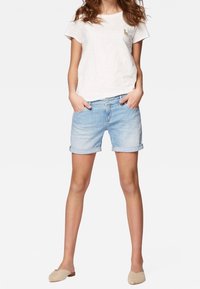 Ljusgrå t-shirt med kort ärm och en liten guldsymbol på bröstet, ihop med uppvikta denimshorts och beige slip-on skor.