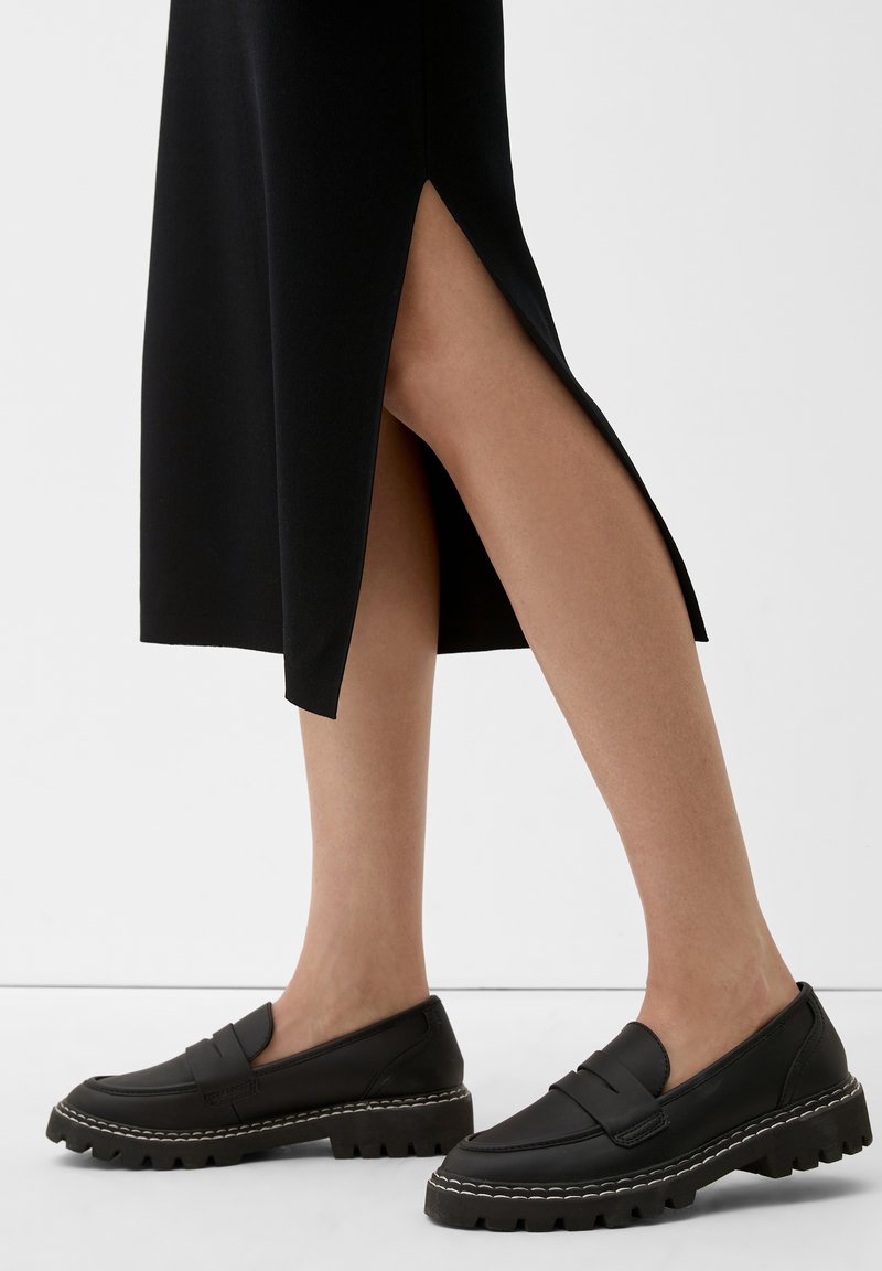 Frau, die schwarze Loafer mit klobigen Sohlen und weißer Naht trägt, kombiniert mit einem schwarzen Rock mit einem seitlichen Schlitz.