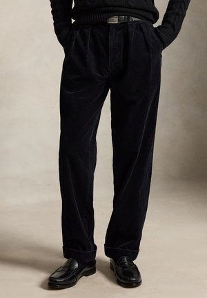 Personne portant un pantalon en velours côtelé noir avec une ceinture, un pull noir et des mocassins en cuir noir, debout sur un fond neutre.