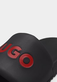 Sandale à glissière noire avec logo "HUGO" rouge en relief sur la large sangle traversant le pied.