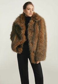 Płaszcz z sztucznego futra w brązowych i czarnych odcieniach, z szerokim kołnierzem i objętością teksturowaną. Noszony na czarnym golfie i spodniach.