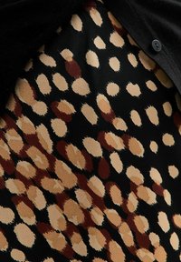 Svart tyg med ett mönster av beige och bruna fläckar, med en mjuk textur och en knappdetalj nära det övre vänstra hörnet.