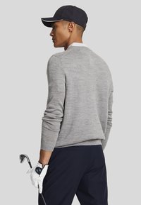 Maglione grigio con polsini e orlo a costine, indossato sopra una camicia bianca con colletto. Pantaloni scuri, tiene un bastone da golf con un guanto bianco. Cappellino nero.