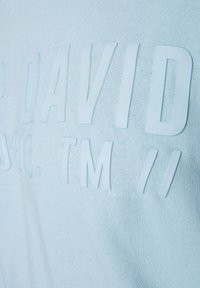 Camiseta de tela azul claro con letras blancas en relieve y textura que dicen "DAVID" y texto adicional, creando un contraste sutil en la superficie.