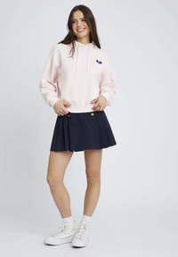Sweat-shirt à capuche rose avec un motif bleu, assorti à une jupe plissée bleu marine et des baskets blanches. Chaussettes blanches avec un détail en dentelle.