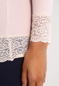 Vaaleanpunainen ribbitekniikalla kudottu paita, jossa on pitsikoristeet hihoissa ja helmassa. Yksityiskohtainen kukkakuvioinen pitsi erottuu sileästä kankaasta.