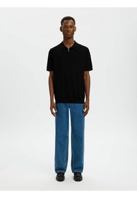 Jeune homme debout portant un polo noir à manches courtes, un jean droit bleu et des chaussures noires, sur un fond uni.