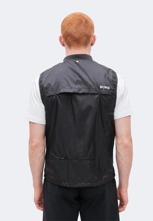 Άτομο με κόκκινα μαλλιά που φοράει μαύρο γιλέκο χωρίς μανίκια πάνω από λευκό πουκάμισο, στέκεται με την πλάτη προς τον θεατή σε απλό φόντο.