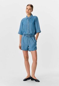 Conjunto de camisa y pantalones cortos de mezclilla azul claro, con cuello, frente abotonado, cintura elástica y mangas enrolladas. El look se completa con calzado negro.
