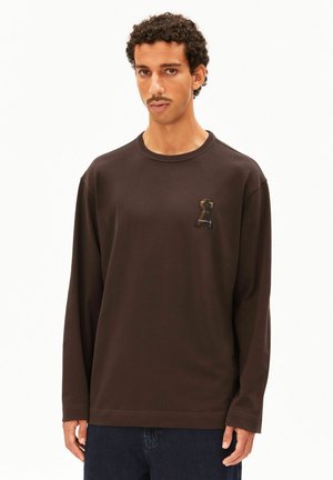 Mann mit kurzen lockigen Haaren und Schnurrbart trägt ein dunkelbraunes langärmeliges Hemd mit einem kleinen bestickten Schlüsselloch-Design auf der Brust.