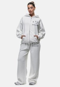 Vrouw in een witte oversized zip-up hoodie en bijpassende brede sweatpants met zwarte tekst, staand met handen in de zakken tegen een witte achtergrond.