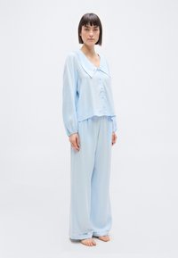 Γυναίκα που στέκεται ξυπόλητη φορώντας ανοιχτό μπλε πυτζάμα με μακριά μανίκια, ευρύ γιακά, πουκάμισο με κουμπιά και παντελόνι με φαρδιά μπατζάκια και ασημένιο περίγραμμα.