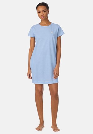 Donna in piedi a piedi nudi che indossa un vestito casual a maniche corte a righe azzurre e bianche con un piccolo logo ricamato sul petto.