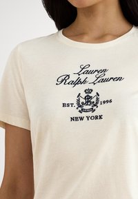 Μπεζ βαμβακερό μπλουζάκι με κέντημα σε σκούρο μπλε λογότυπο με τα γράμματα "Lauren Ralph Lauren" και "New York", ίδρυση 1996.