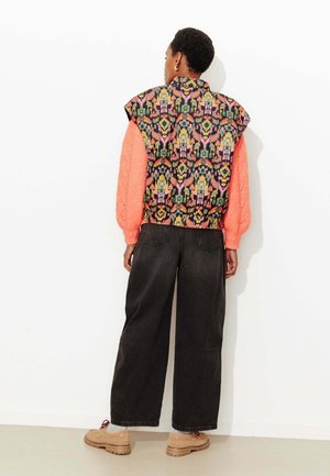 Veste à motifs colorés sur fond noir avec des détails roses, associée à un pull en maille orange surdimensionné et un pantalon noir ample.