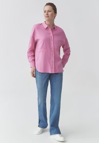 Donna in piedi con una mano in tasca, indossa una camicia button-up rosa morbida, jeans blu con spacchi laterali e sneakers bianche.