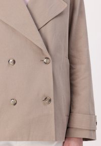 Beige dvouřadá bunda s velkými klopami, texturovaná tkanina a dvě výrazné knoflíky na přední straně a manžetách.