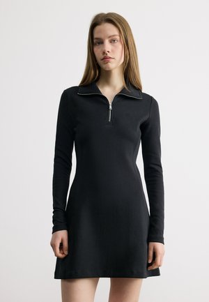 Fiatal nő, aki egy fekete, testhez simuló, hosszú ujjú ruhát visel, amely elején cipzáros gallérral rendelkezik, és egy egyszerű, világos háttér előtt áll.