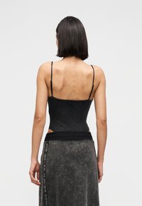 Γυναίκα με σκούρα μαλλιά μέχρι τους ώμους, φοράει μαύρο αμάνικο μπλουζάκι και σκούρο ξεβαμμένο φόρεμα, στέκεται με την πλάτη προς τα εμπρός.