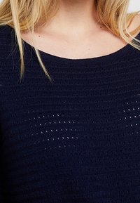 Granatowy sweter dzianinowy z szerokim dekoltem i fakturowanym wzorem z małymi, otwartymi oczkami. Miękka, lekka tkanina.