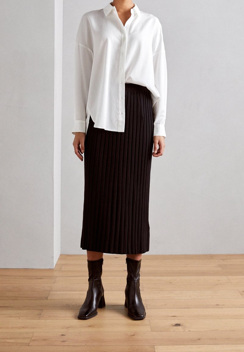 Bílá košile s dlouhými rukávy a knoflíky v kombinaci s černou midi sukni s plisováním a černými kotníkovými botami. Hladká tkanina a přesný střih.