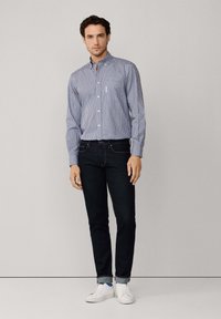 Hombre con camisa de cuadros azul y blanca, jeans oscuros arremangados y zapatillas blancas, de pie contra un fondo liso.