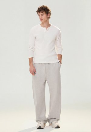 Mladi moški v beli dolgoročni henley majici, svetlo sivih širokih hlačah in belih supergah, stoji z rokami v žepih.