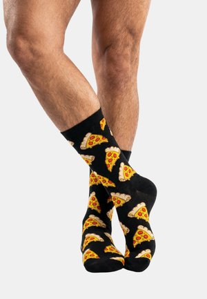 Nogi skrzyżowane, ubrane w czarne skarpetki z wzorem żółto-czerwonych kawałków pizzy na jednolitym tle.