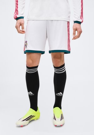Nogi w białych krótkich spodenkach piłkarskich z logo Realu Madryt, długie czarne skarpety Adidas i jaskrawo żółte korki stojące na białym tle.