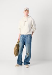 Beige Kapuzenpullover mit einer Fronttasche, hellblaue Jeans mit einem aufgerissenen Knie, beige Kappe und beigefarbener Rucksack, kombiniert mit hellen Schuhen.