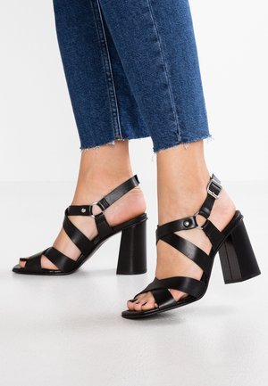 Sandales à talons noirs à bout ouvert, avec des straps croisées et une fermeture à boucle, reposant sur un talon carré avec une finition lisse.
