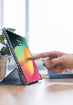 Finger touching screen of tablet in a standing case on a wooden desk with blurred background and plant nearby.