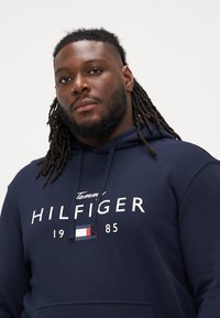 Μπλούζα φούτερ ναυτικού μπλε χρώματος από μαλακό υλικό, με άσπρη εκτύπωση λογότυπου, ρύθμιση με κορδόνι στην κουκούλα και τσέπη καγκουρό. Κλασικός casual σχεδιασμός.