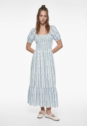 Kvinde står med hænderne bag ryggen iført en hvid midi-kjole med blå blomstrede lodrette striber og puffede korte ærmer.