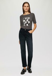 Grijze T-shirt met grafische print, zwarte rechtgesneden jeans en glanzende zwarte instappers. Model staat tegen een neutrale achtergrond.
