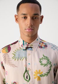 Short-sleeve, collared shirt in pastel pink with vibrant floral, leafy, and abstract patterns; features a button-up front and textured fabric.