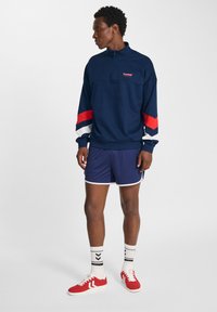 Maglione navy con zip a un quarto, con accenti a strisce rosse e bianche sulle maniche, abbinato a pantaloni corti navy e scarpe da ginnastica rosse.