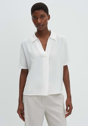 Frau mit kurzen Haaren, die eine locker sitzende weiße Kurzarmbluse und hellbeige Hose trägt, vor einem einfarbigen Hintergrund stehend.