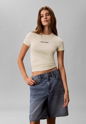 Jeune femme debout portant un t-shirt crème ajusté Calvin Klein et un short en jean ample, taille haute, longueur genou, sur fond gris.