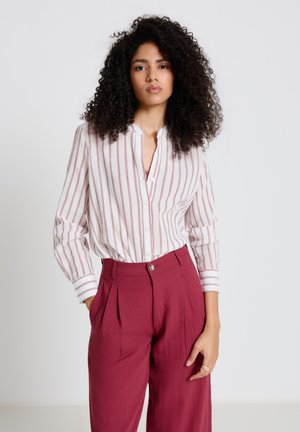 Femme aux cheveux bouclés portant un chemisier blanc à rayures rouges et un pantalon rouge taille haute, debout devant un fond clair uni.