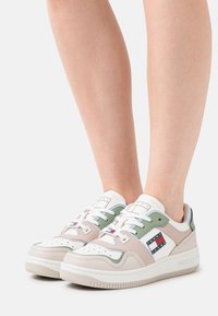 Baskets blanches et pastel avec une tige en cuir, présentant des accents verts et beiges, une boîte à orteils perforée, et un patch logo sur le côté.