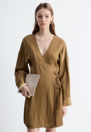 Joven mujer con un vestido cruzado de color marrón claro que sostiene un bolso de mano beige texturizado, complementado con pendientes de aro dorados y múltiples pulseras.