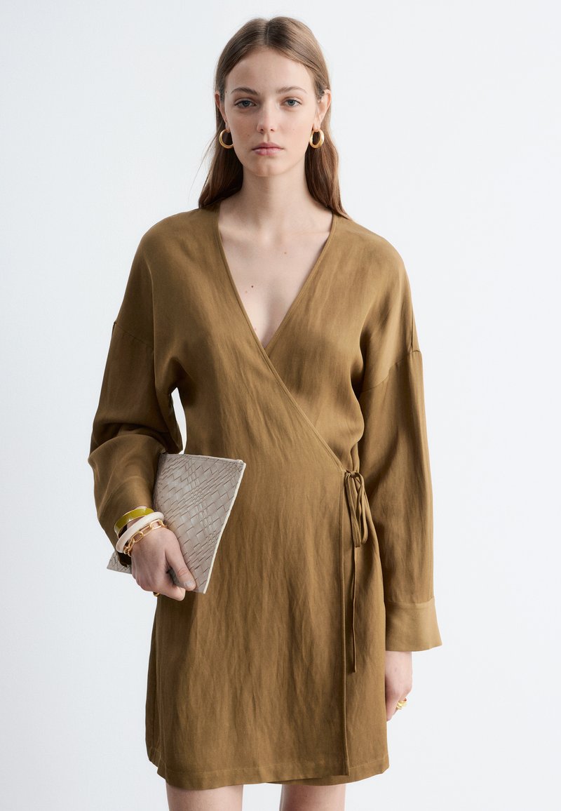 Jeune femme portant une robe portefeuille marron clair, tenant une pochette beige texturée, accessoirisée avec des boucles d’oreilles créoles dorées et plusieurs bracelets.