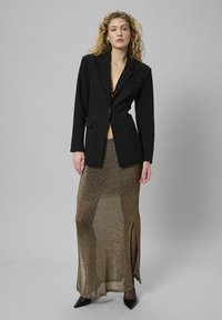 Schwarzer maßgeschneiderter Blazer, langer metallisch goldener Rock mit Seitenschlitz, Riemchenschuhe. Glatte Stofftextur mit einem schimmernden Effekt.