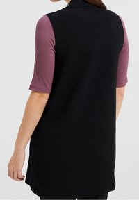 Svart ärmlös tunika med hög krage, kombinerad med en ribbad mauve topp med kort ärm. Slät textur, sidoflikar och avslappnad passform.