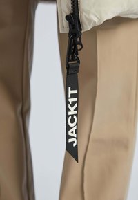Tirette de fermeture éclair en nylon noir avec le texte blanc "JACK1T", attachée à un tissu pâle, à côté d'un pantalon en toile lisse de couleur beige.